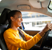 A woman in a yellow shirt drives a car and smiles broadly.